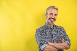 A middle-aged man with gray hair and a beard smiles with arms crossed, standing in front of a bright yellow background. His patterned button-up shirt adds style to this portrait by a corporate headshot photographer Manchester.