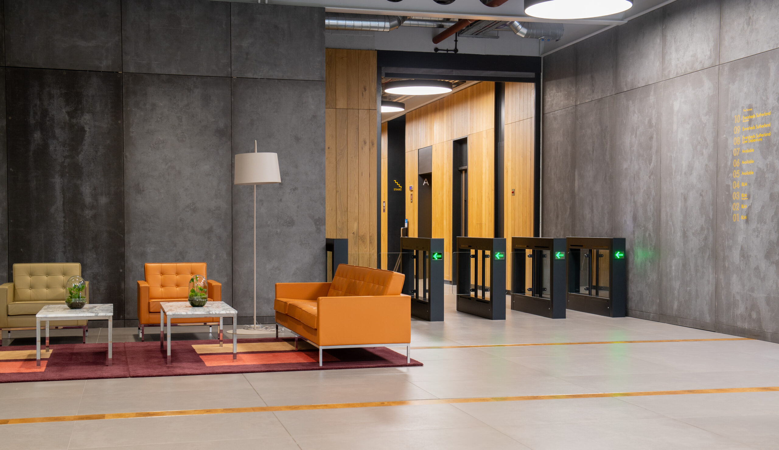 A modern office lobby with gray walls, orange and beige sofas, small glass tables, and potted plants. Security turnstiles with green lights are in the background near wooden wall panels. 2 New Bailey Square Manchester