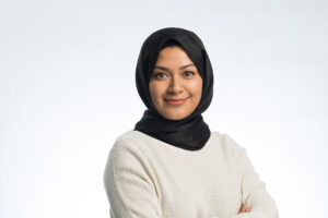 A woman wearing a black hijab and a white sweater smiles confidently with her arms crossed against a plain light background, exemplifying How to take a professional headshot Manchester style.