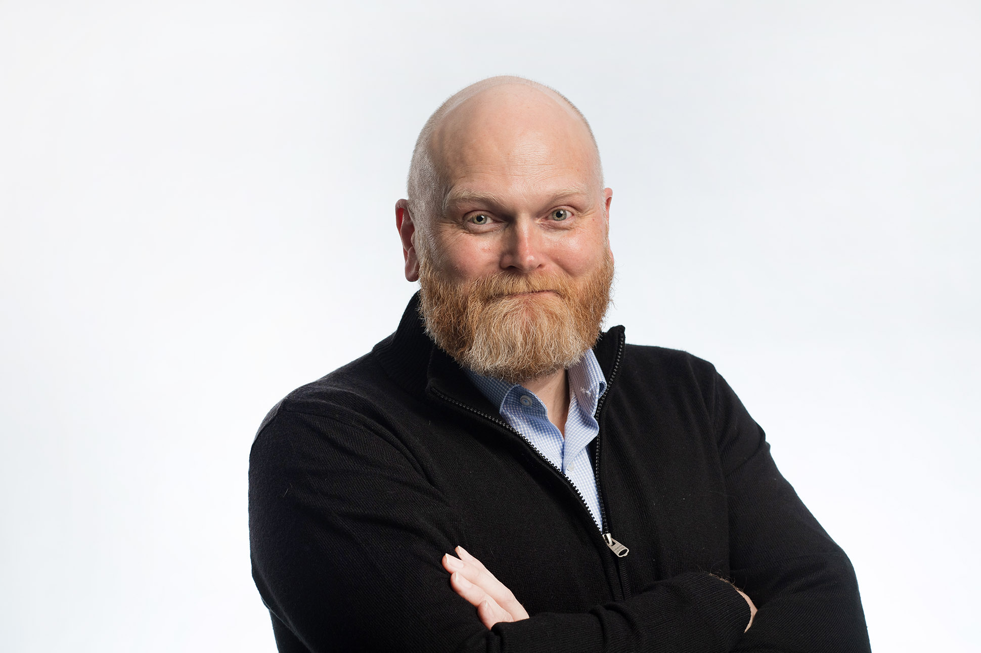 A bald man with a reddish beard, wearing a black zip-up sweater over a light blue shirt, stands with arms crossed and smiles at the camera against a plain white background—showcasing how to take a professional headshot in Manchester.
