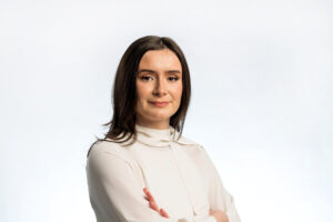 A woman with long dark hair wearing a white blouse stands with arms crossed, smiling confidently against a plain white background—showcasing an example of how to take a professional headshot in Manchester.