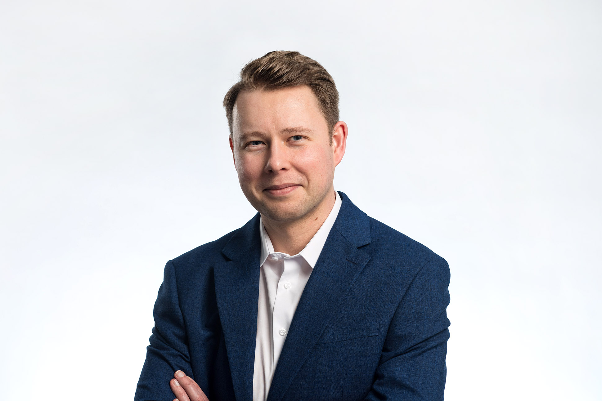 A man with light brown hair, wearing a navy blue suit and white shirt, stands against a plain white background, smiling softly with his arms crossed—an ideal example of how to take a professional headshot in Manchester. Business headshot portrait photographer.