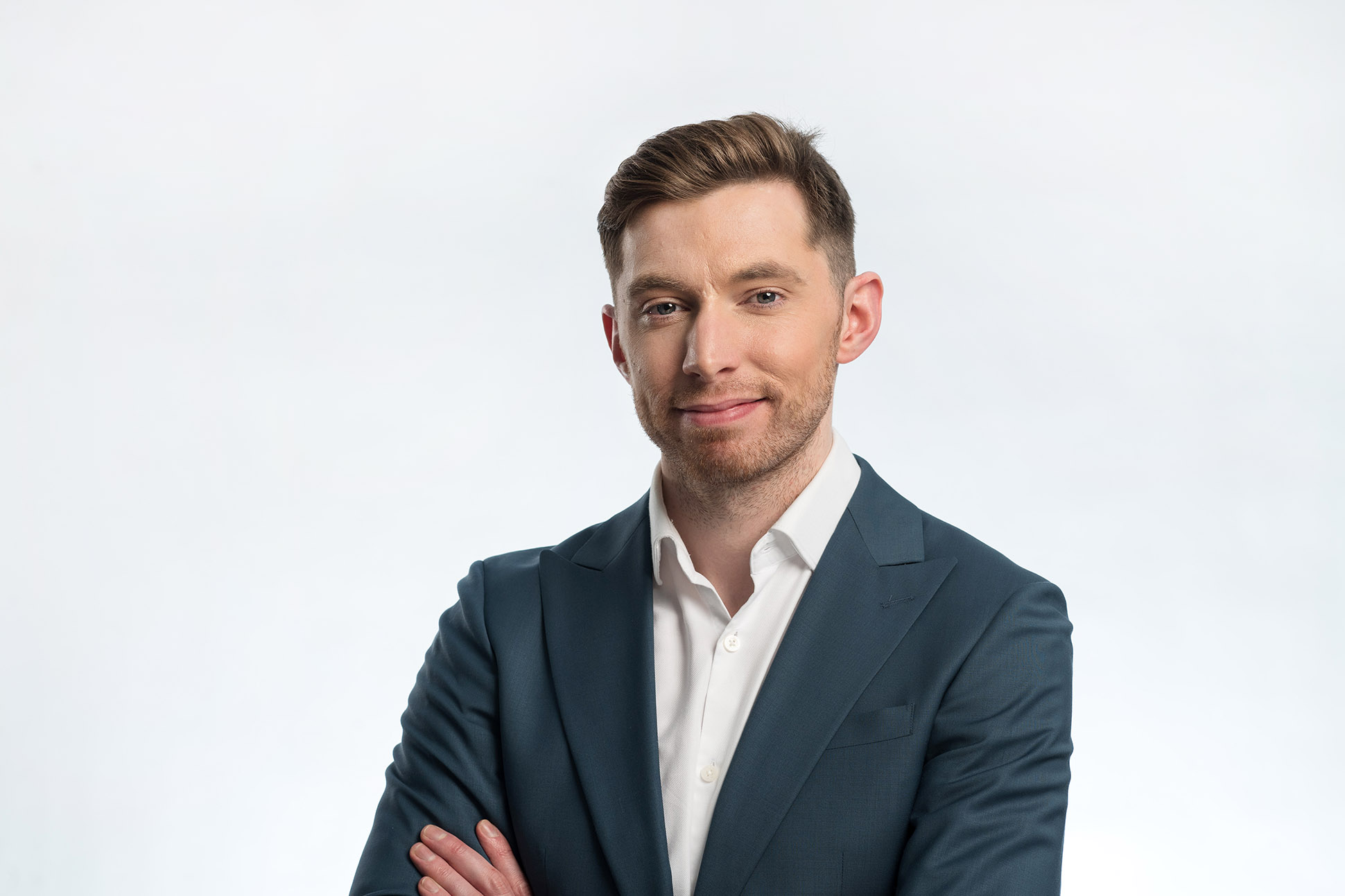 A man in a blue suit and white shirt stands with his arms crossed, smiling slightly, against a plain white background—a good example of how to take a professional headshot in UK.