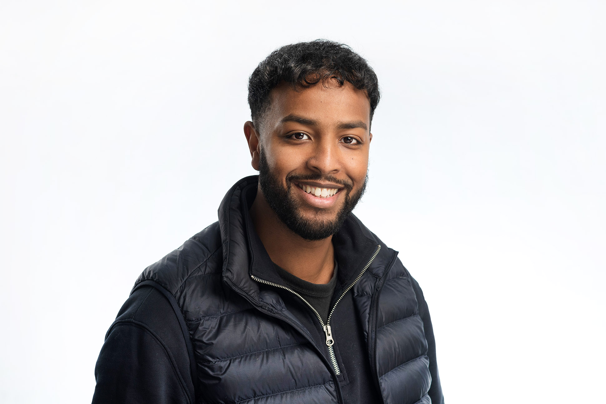 A young man with short curly hair and a beard smiles at the camera, dressed in a black quilted vest over a black shirt. This image is a great example of how to take a professional headshot in Manchester styled against a plain white background.