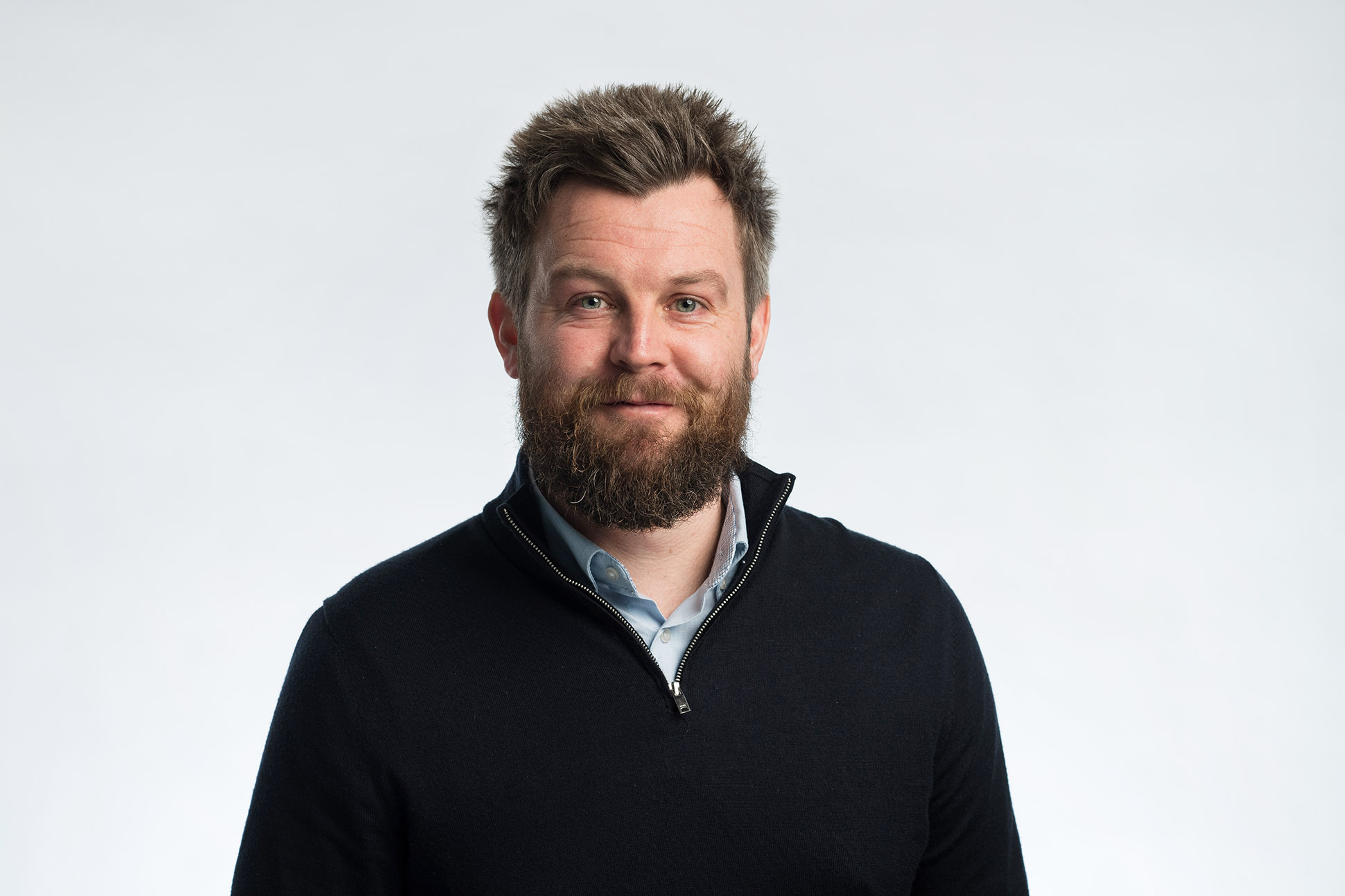A man with a beard and short, spiked hair is smiling slightly in this professional headshot—an example of how to take a professional headshot - Manchester style. He wears a dark sweater over a collared shirt; the background is plain and light-colored. Relaxed headshot photography throughout the UK.