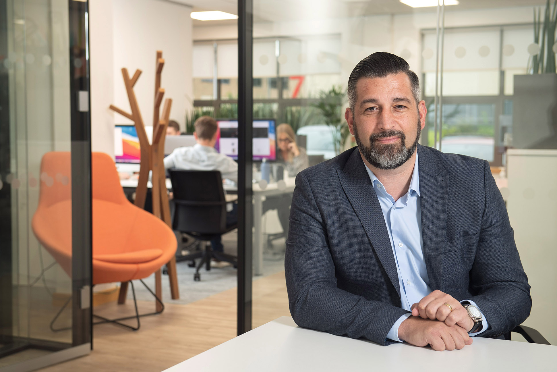 A man with dark hair and a beard in a blue suit sits at a desk in a modern office. Behind him, people work at computers. The office has glass walls, an orange chair, and modern decor. relaxed corporate headshot photographer thoughout the UK