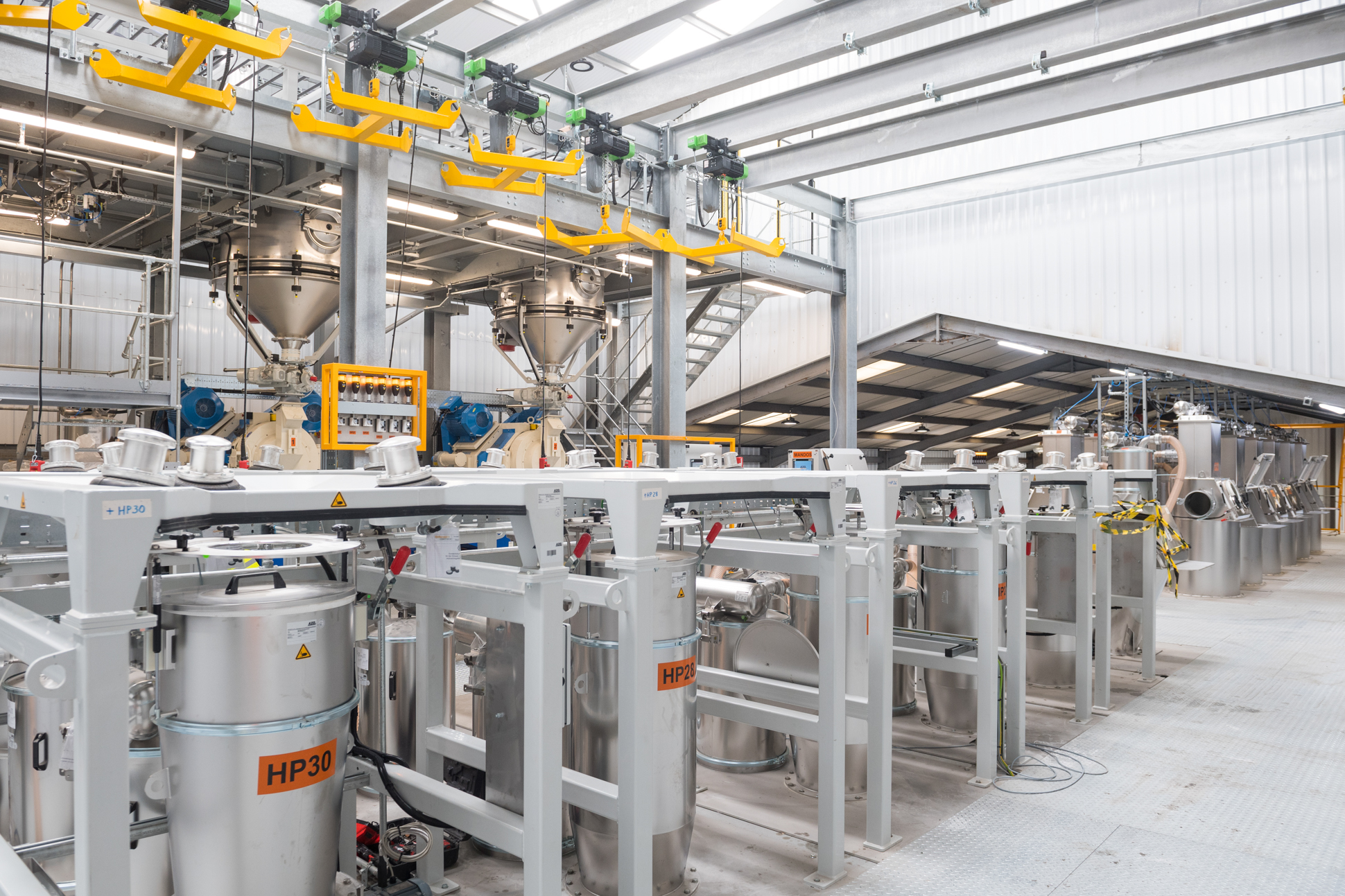 A modern industrial facility with large metal machinery, storage tanks, and pipes, bright lighting, and a clean, organized layout. Overhead rails and platforms are visible, with equipment labeled HP30. Industrial factory equipment photography.