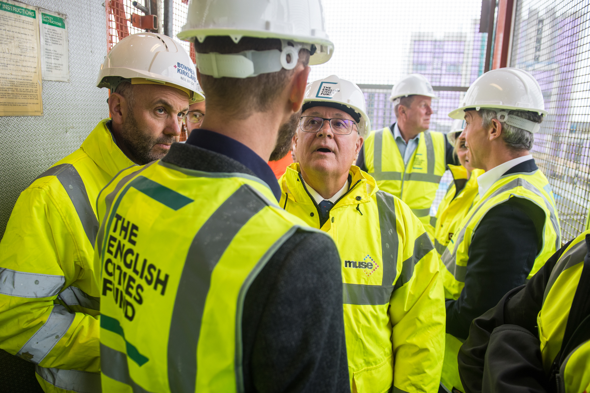 A group of people wearing hard hats and high-visibility jackets stand together on a construction site, engaged in conversation. One jacket reads The English Cities Fund. Some individuals are looking attentively at someone speaking. Construction site tour