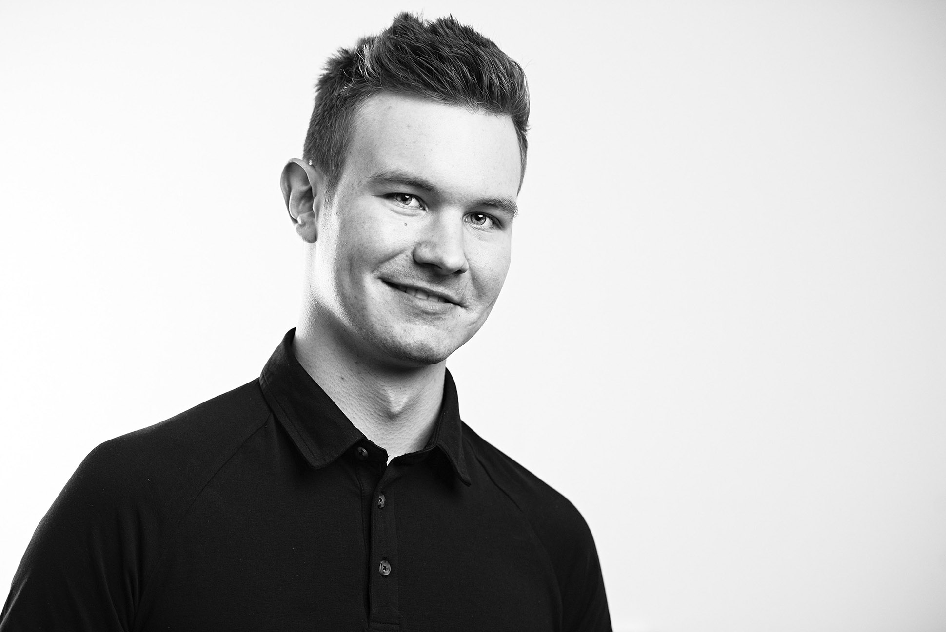 Black and white photo of a young man with short hair, wearing a dark collared shirt, smiling slightly, and looking at the camera. The background is plain and light-coloured. Good example of headshot photography in Manchester