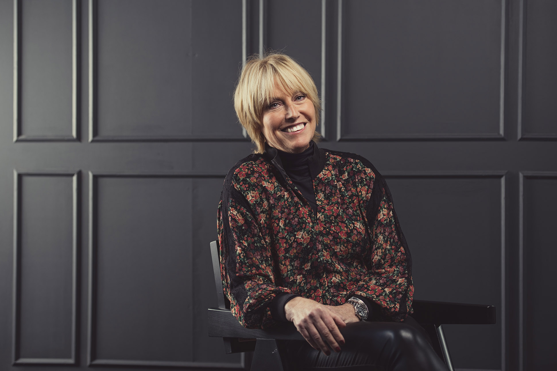 A woman with short blonde hair, wearing a floral-patterned top and black pants, sits on a chair and smiles in front of a dark, paneled wall—an ideal example for anyone seeking tips on how to take a professional headshot in Manchester. Modern professional headshot portrait photographer.