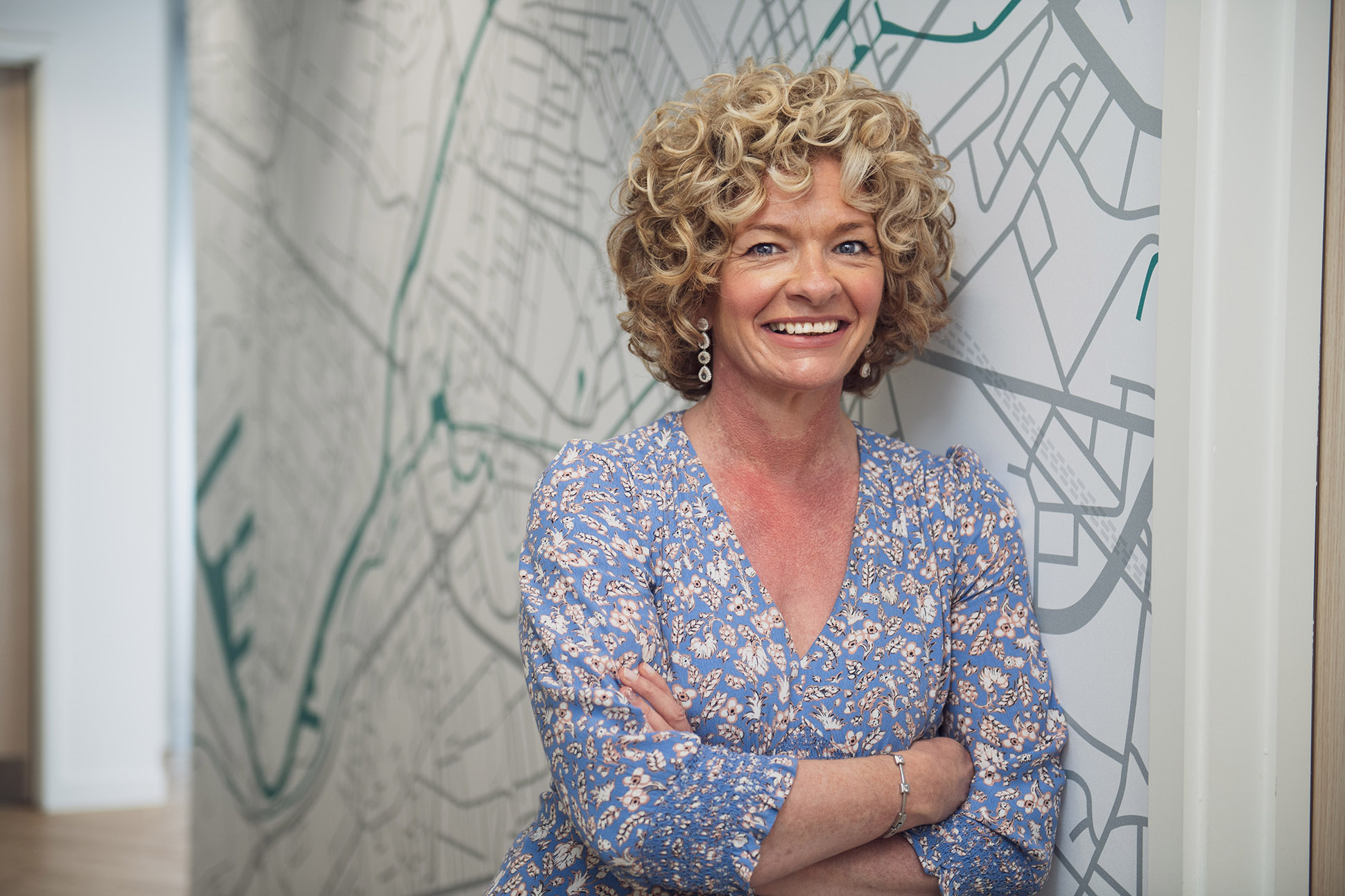 A woman with curly blonde hair, wearing a blue patterned dress, stands smiling with arms crossed in front of a wall decorated with a map design—ideal inspiration if you're wondering how to take a professional portrait or headshot in Manchester.