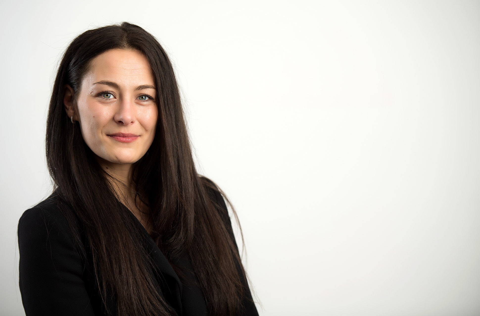 A woman with long dark hair and a black blazer stands against a plain white background, smiling softly at the camera. Jon Parker Lee headshot photography throughout the UK.