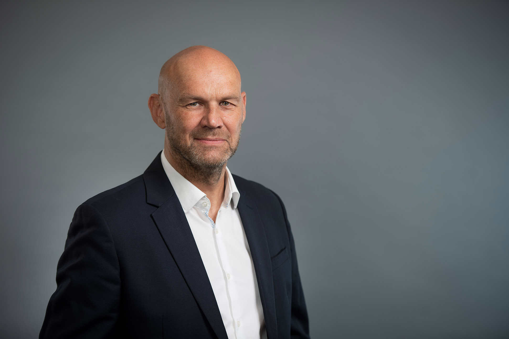 Bald man with a beard wearing a dark suit and white shirt, standing in front of a plain gray background, looking at the camera with a slight smile. Corporate headshot photographer Manchester.
