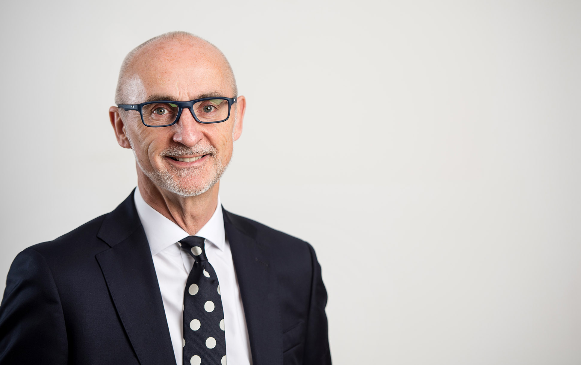 A smiling older man with glasses, wearing a navy suit, white shirt, and polka dot tie, stands against a plain white background. Freelance professional head shot portrait photography