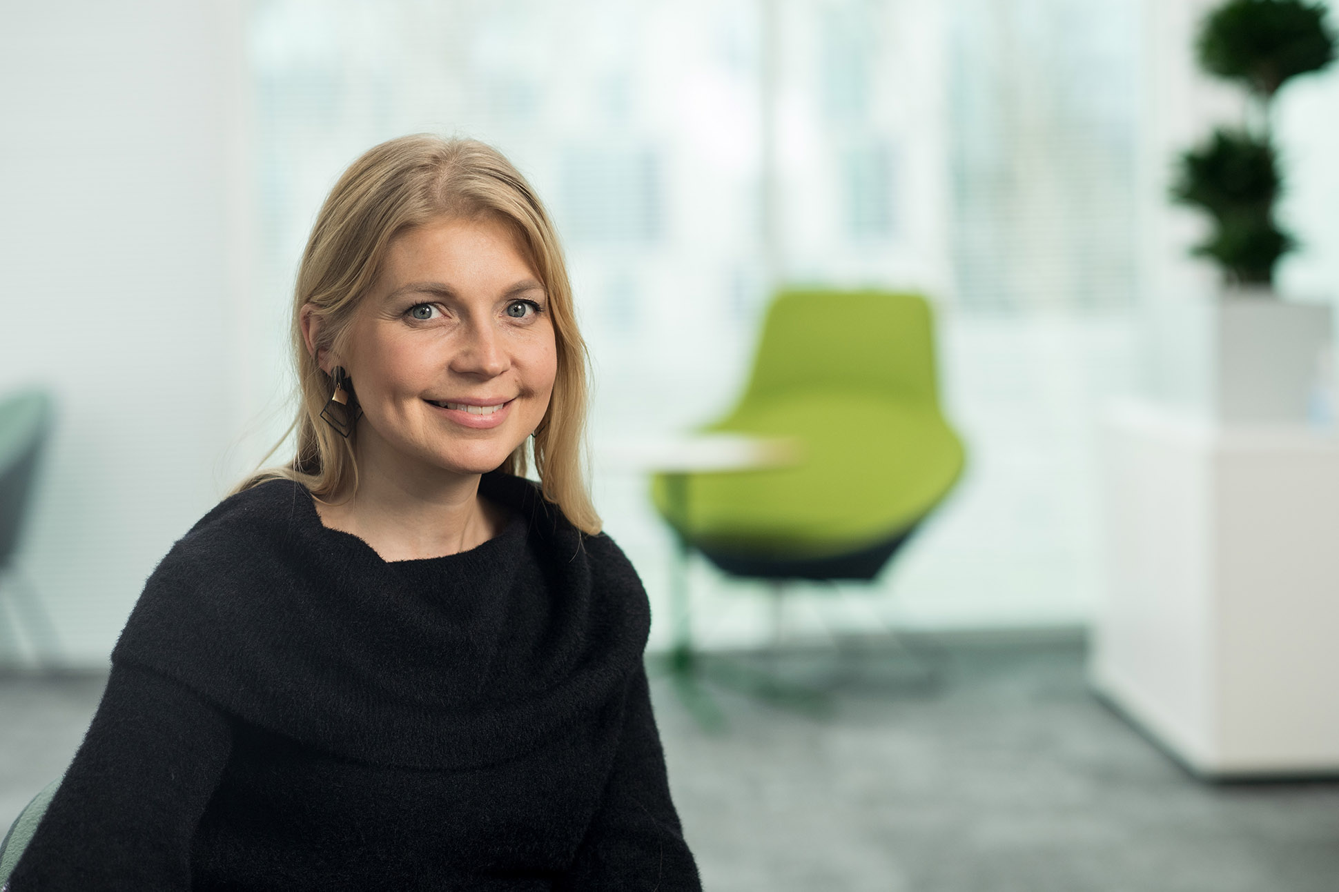 A woman with blonde hair wearing a black sweater smiles while sitting in a modern office space—an inviting example of how to take a professional headshot -Manchester, featuring neutral tones and a green chair in the background.
