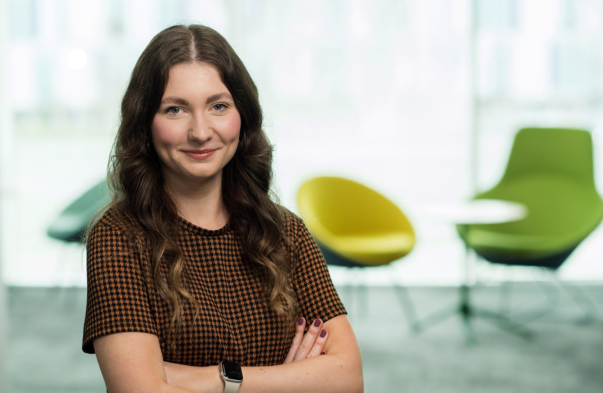 A woman with long, wavy brown hair and a checked shirt stands smiling with arms crossed in a modern office posing for her hreadshot. Blurred green and yellow chairs and a round table are visible in the background. Portrait photographer at law firm Manchester.