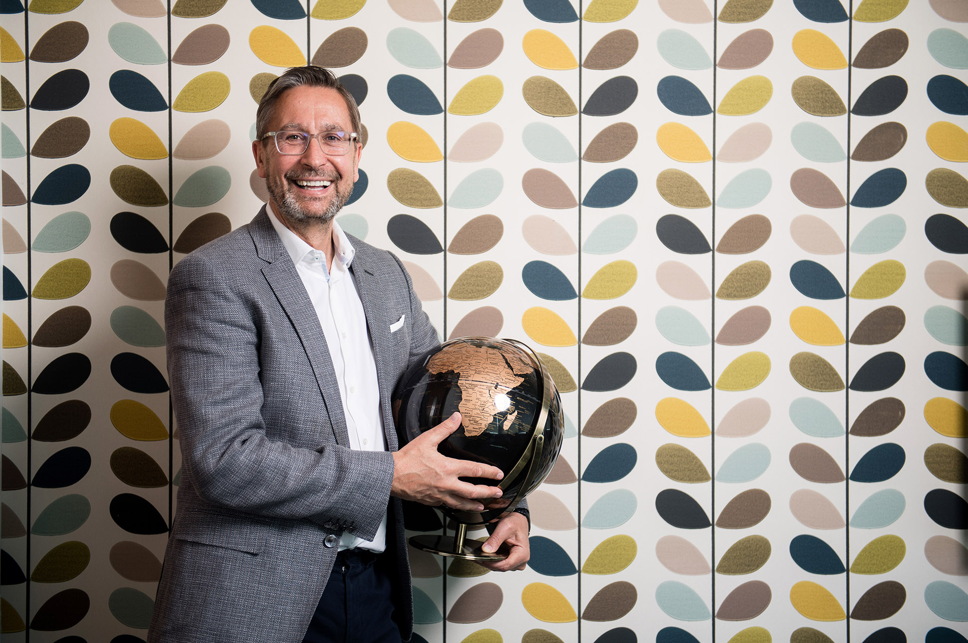 A smiling man in a gray suit holds a black and gold globe, standing in front of a colorful, patterned wall with leaf designs. Travel Counsellers commercial photography Manchester