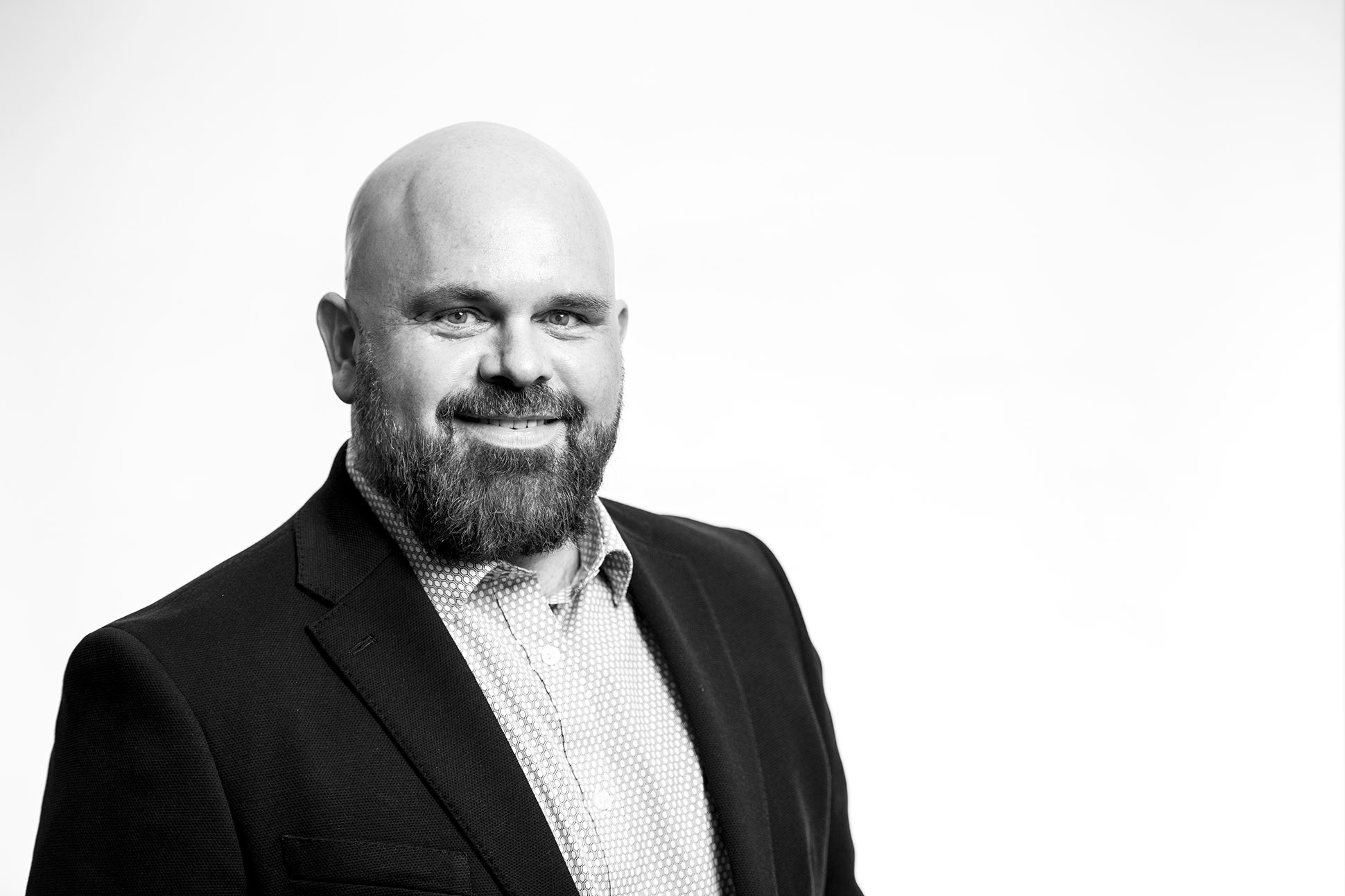 A bearded, bald man wearing a blazer and collared shirt smiles at the camera in a black-and-white portrait with a plain white background, capturing the essence of how to take a professional headshot. Head shots for Manchester staff operating throughout the country.