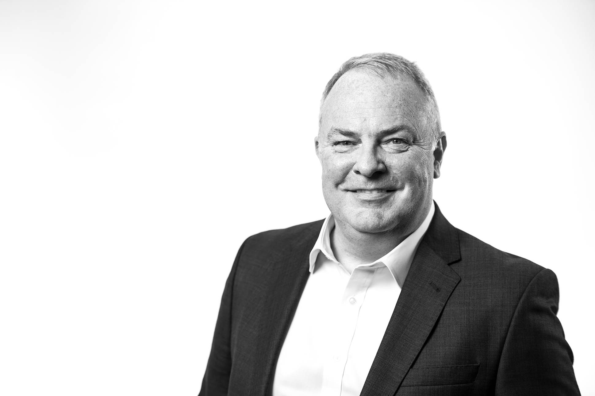 Black and white portrait of a middle-aged man in a suit jacket and white shirt, smiling, against a plain white background. Headshot photographer can visit all your regional offices.