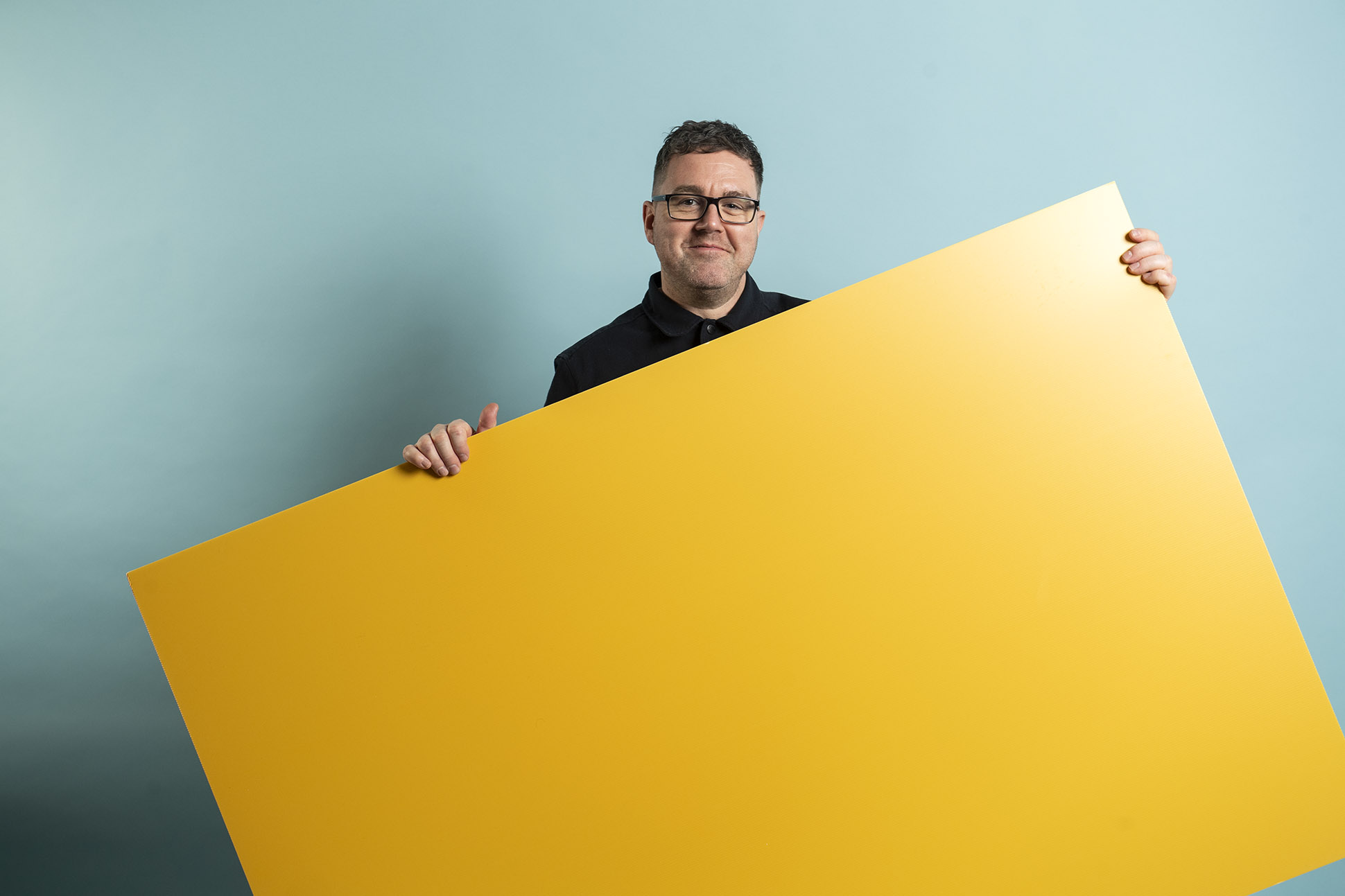 A man wearing glasses and a black shirt stands against a light blue background, holding a large rectangular yellow board in front of him.