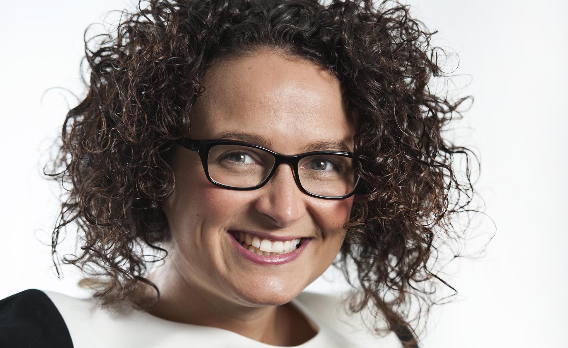 A woman with curly dark hair and glasses smiles at the camera. She is wearing a white top with a black collar and has a light background behind her. Relaxed corporate headshot at regional offices throughout the UK.