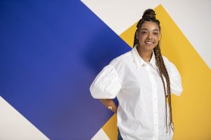 A woman with long braids, wearing a white blouse with puffy sleeves, stands smiling in front of a geometric blue and yellow background—perfect for a corporate headshot photographer Manchester portfolio.