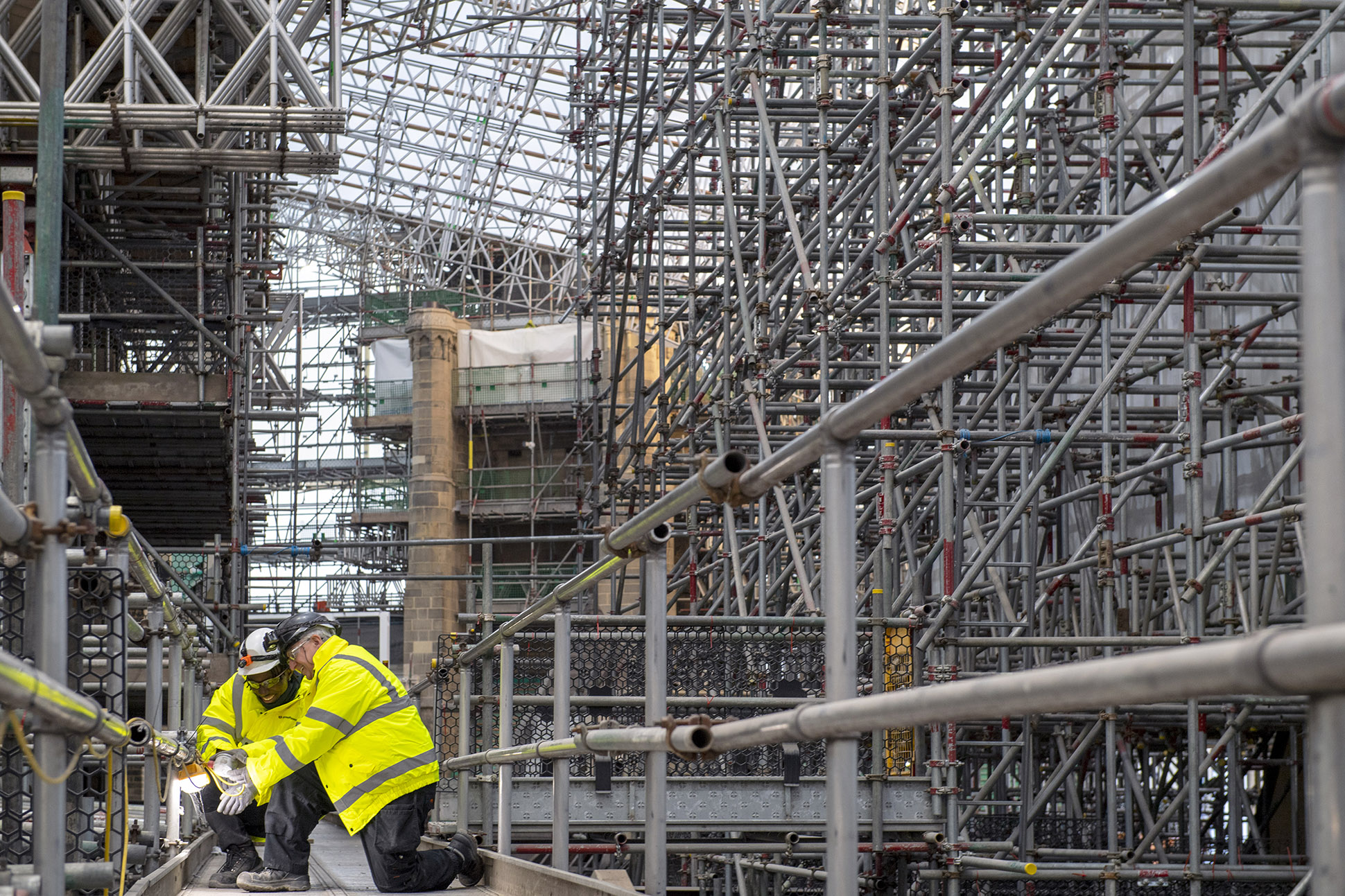 Two construction workers in yellow safety jackets and helmets kneel on scaffolding surrounded by a complex network of metal pipes and beams at a construction site. Industrial Building photography