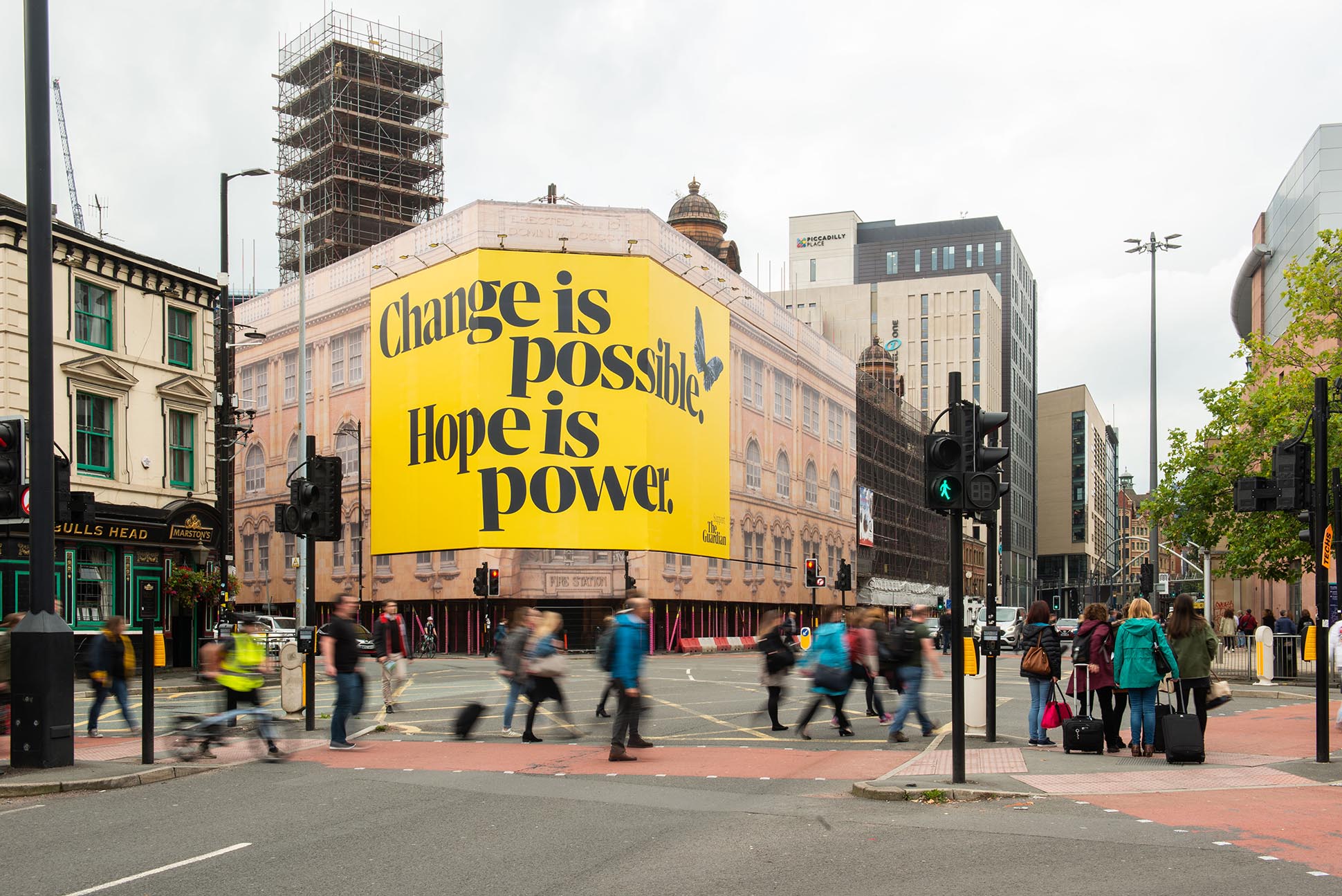 A busy city street intersection with people crossing. A large yellow billboard on a building reads, Change is possible. Hope is power. Surrounding buildings and traffic lights are also visible. OOH photography Piccadilly Manchester