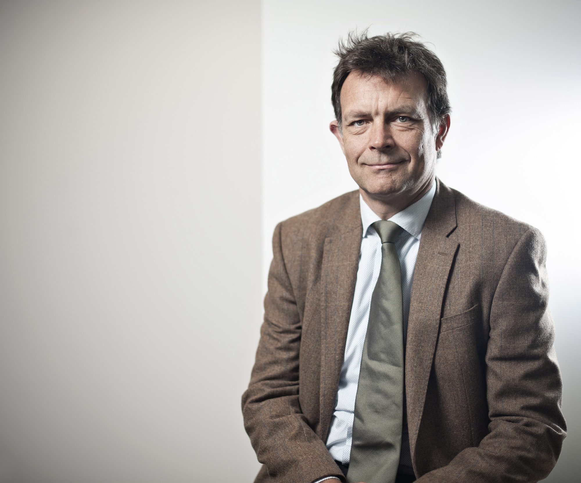 A middle-aged man wearing a brown blazer, light blue shirt, and green tie sits against a plain light-colored background, looking at the camera with a slight smile. Business portrait photography