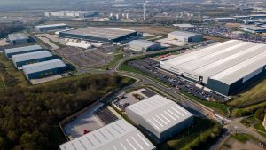 Aerial view by a skilled drone photographer and videographer Manchester of a modern industrial park with large warehouses, parking lots, access roads, and green spaces, all surrounded by buildings and open land on a clear day. Large logistics centre aerial photograph from drone.