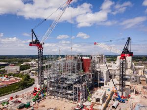 A large construction site in Manchester with two cranes assembling a steel-framed building under a partly cloudy sky, captured by a drone photographer and videographer, surrounded by equipment, materials, and partially built structures. Drone video of large industrial site.