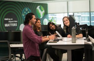 Four colleagues stand and sit around a desk in a modern office, smiling and collaborating. Captured by a corporate photographer Manchester, the scene features a green "Making an impact that MATTERS" sign, computer monitors, and large windows. Business team photography