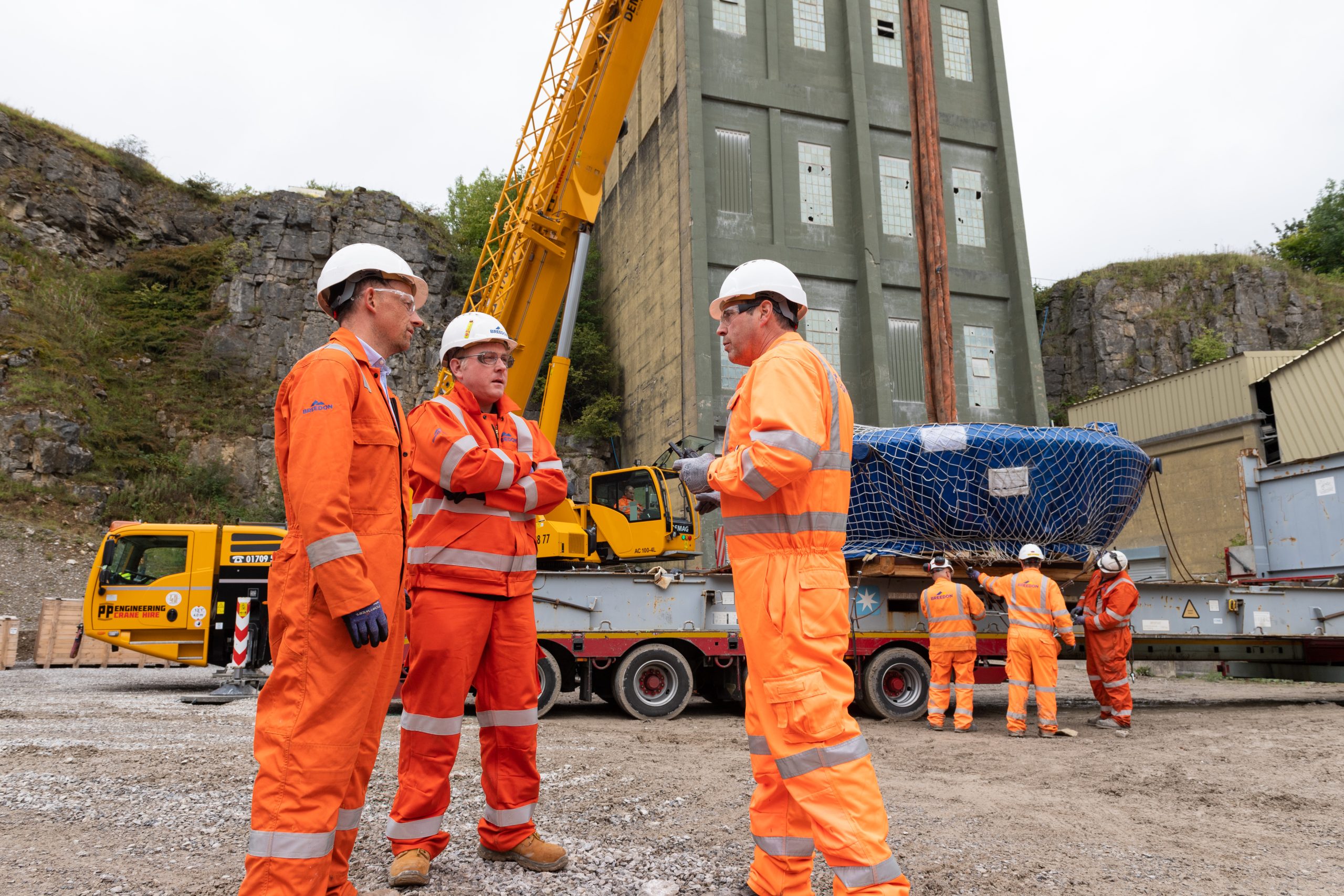 Three construction workers in orange safety gear and white helmets talk near a building site, as seen in this industrial installation film Sheffield, with a crane, a large building, and more workers handling equipment in the background.